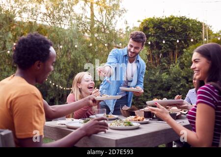 Multi-Cultural Friends At Home Sitting At Table Enjoying Food At Summer Garden Party Stock Photo