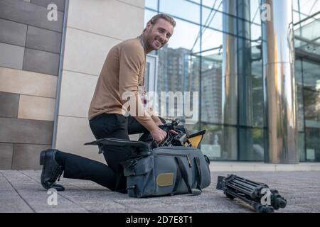 Smiling cameraman standing on one knee outside, taking equipment out of case Stock Photo