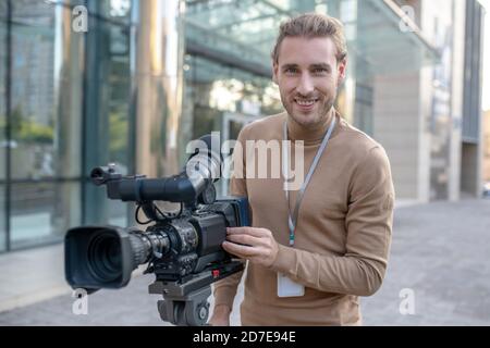 Fair-haired cameraman standing behind camera in front of modern building Stock Photo