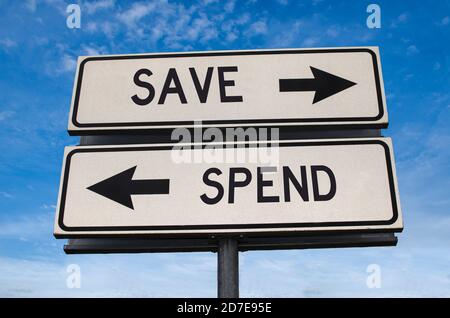 Save vs spend. White two street signs with arrow on metal pole with word. Directional road. Crossroads Road Sign, Two Arrow. Blue sky background. Stock Photo