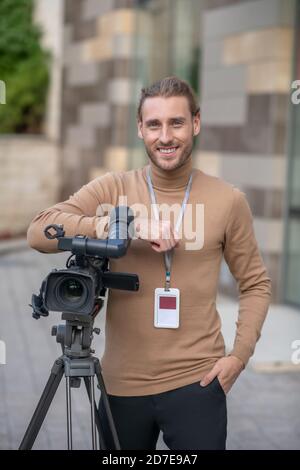 Smiling fair-haired cameraman standing next to camera, leaning on elbow Stock Photo