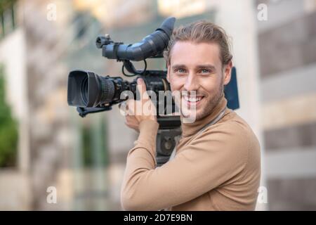 Smiling fair-haired operator standing with camera on his shoulder outside Stock Photo