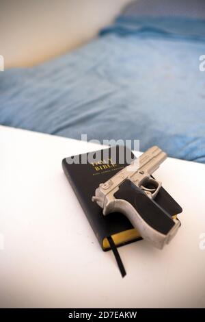 a silver replica berretta 9mm pistol on a black leather Holy Bible rests on a white desk next to a made bed in a bedroom in a home Stock Photo