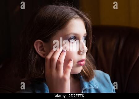 A natural portrait of a tween or teen girl with hand on face sat in an armchair, looking contemplative Stock Photo