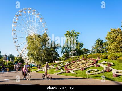 Cyclists passing by L'Horloge Fleurie, the flower clock in Geneva, with the Ferris wheel on the wharf of the Lake Geneva on a sunny summer day. Stock Photo