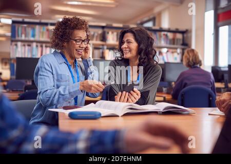 Teacher With Mature Female Adult Student Using Mobile Phone At Table Working In College Library Stock Photo