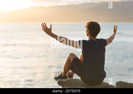 Happy young man feeling good sitting on top of a mountain cliff facing the ocean view and raising arms up to the sky. Hope and gratitude. Stock Photo