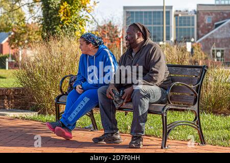 Frederick, MD, USA 10/14/2020: An interracial couple (caucasian woman and African American man) are sitting together on a park bench. The woman wears Stock Photo