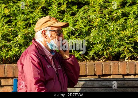 Frederick, MD, USA 10/14/2020: An elderly man with baseball hat is sitting on a bench and talking on phone. He has removed his face mask, he wears due Stock Photo