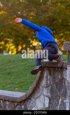 A hyperactive kid is jumping off a high wall as he is running away. The courageous boy wears track pants, sneakers and a hooded coat. He is full of en Stock Photo