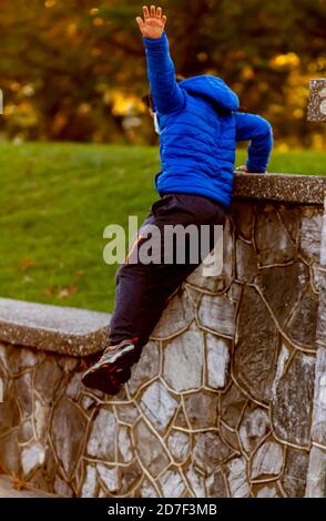 A hyperactive kid is jumping off a high wall as he is running away. The courageous boy wears track pants, sneakers and a hooded coat. He is full of en Stock Photo