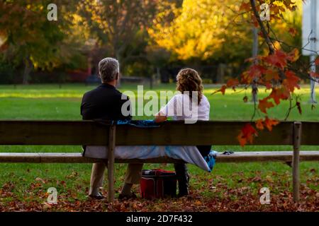 A middle aged caucasian couple is sitting on a wooden bench in a park covered with fallen leaves at sunset. A scenic autumn concept with romance and t Stock Photo