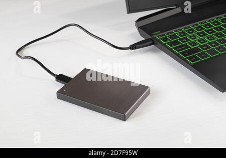 An external hard drive is connected to the laptop for transferring information on a white wooden table. Horizontal orientation, selective focus. Stock Photo