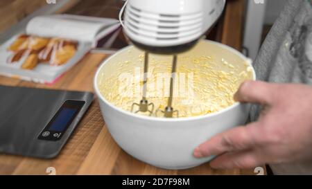A man holds an electric hand mixer. He mixes butter, eggs and sugar. A short pastry is prepared. Close up from the kitchen. Stock Photo