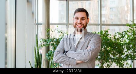 Smiling Caucasian man standing near window in home greenhouse with plants or modern office. Portrait of successful happy bearded young man. Handsome Stock Photo