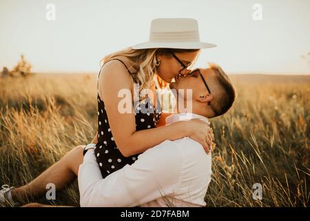 A girl in hat sits in hands guy on ground in middle of tall grass in meadow Stock Photo
