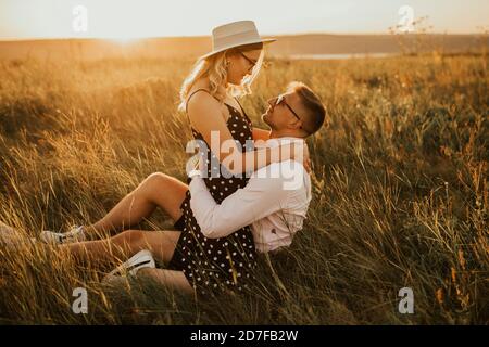 A girl in hat sits in hands guy on ground in middle of tall grass in meadow Stock Photo