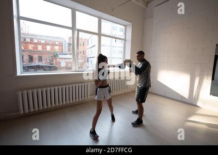 A man with boxing paws on his hands teaches the technique of hitting a novice girl in a light gym Stock Photo
