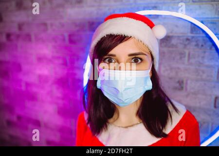 Young woman in medical mask and Santa Claus suit against illuminated wall. Close up of brunette in Christmas hat and protective mask. Concept of safe Stock Photo