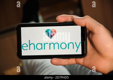 Eharmony logo on the screen of mobile phone in mans hand. Application for dating and acquaintance. Man is laying on the sofa and looking for partner Stock Photo