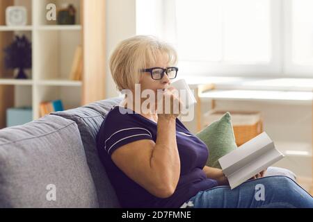Mature woman in glasses relaxing on couch at home, drinking tea and reading a book alone Stock Photo