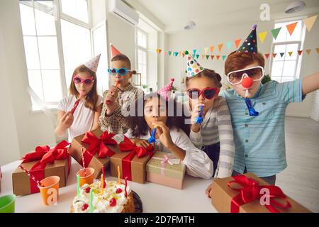 Bunch of happy diverse kids in sunglasses and cone hats blowing noisemakers at birthday party Stock Photo