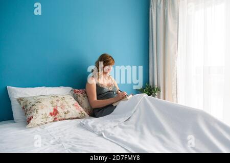 A pregnant smiling woman in a semi-sitting position in her bed while writing something on a notebook. Stock Photo