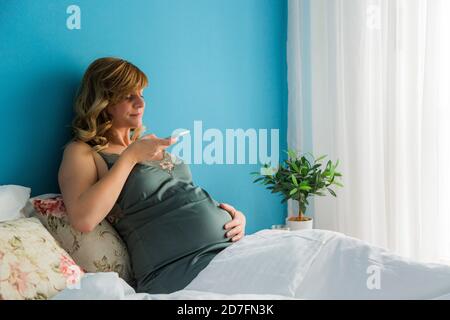 A pregnant woman in a semi-sitting position in her bed while taking photos of her belly with her smartphone. Stock Photo