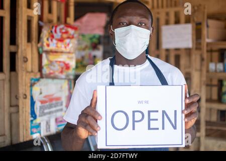 african business owner wearing a face mask and holding an open sign in front of his shop Stock Photo