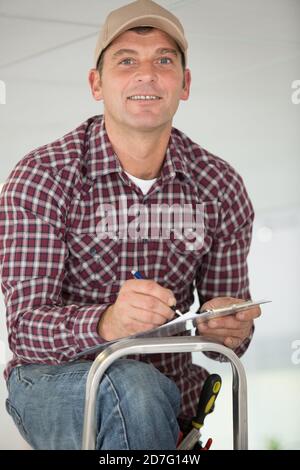mature tradesman on stepladder writing on clipboard Stock Photo
