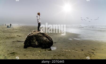 Senior Woman standing on a large Driftwood Log looking out into the Dense Fog over the Pacific Ocean in Cox Bay at the Pacific Rim National Park Stock Photo