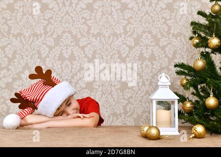 Smiling with his eyes closed, a boy lies on the table on folded arms near the Christmas tree. Stock Photo