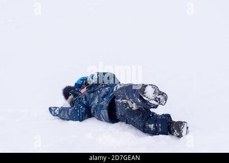 A boy in winter blue clothes lies on the white snow. Outdoor games in the snow. Danger of getting sick, freezing in the snow Stock Photo