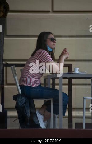 italian girl sitting alone, with a mask held under her chin, at a coffee table enjoying a coffee and reflecting thoughtfully Stock Photo