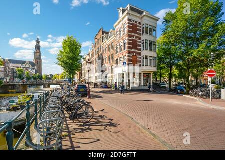 Bicycles parked along a canal near the Westerkerk Church as tourists enjoy the shops and bridges in the historic center of Amsterdam Netherlands Stock Photo
