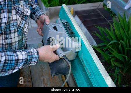 Landscaping of the garden. handyman sharpens a wooden bench on the street in the garden. master removes a layer of dry paint from the wood surface Stock Photo