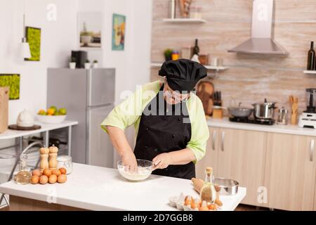 Woman mixing eggs with flour to make dough after traditional recipe on kitchen table. Retired elderly chef with uniform sprinkling, sieving sifting raw ingredients and mixing . Stock Photo