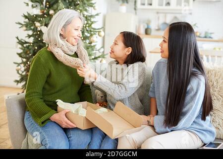 Two daughters giving warm handmade scarf for his mother on Christmas Day they sitting on sofa in the room Stock Photo