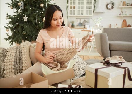Young Asian woman sitting on sofa and getting the warm scarf out of the Christmas present during Christmas Eve Stock Photo