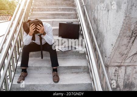 Unemployed stressed young Asian business man with laptop in suit covering face with hands sitting on the stairs. Failure and layoff concept to the dis Stock Photo