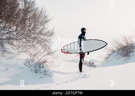 Snowy winter and surfer with surfboard. Winter beach and surfer in wetsuit. Stock Photo