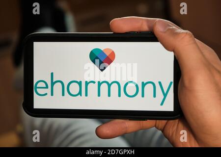 Eharmony logo on the screen of mobile phone in mans hand. Application for dating and acquaintance. Man is laying on the sofa and looking for partner Stock Photo