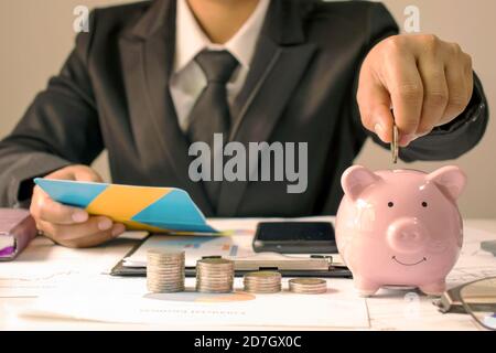 Businessman holds a coin on a piggy bank and is examining a business investment money-saving idea account. Stock Photo
