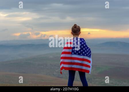 Young little teenager blonde girl holding flag of America USA at sunset outdoors.