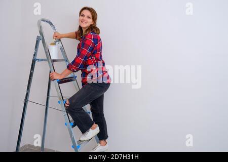 A painter woman stands on a stepladder with a brush and a can of paint, copy space. Painting the walls in a new apartment Stock Photo