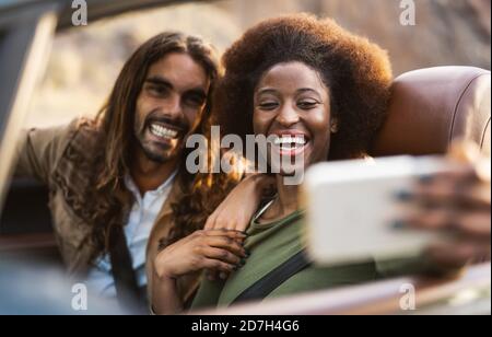 Happy young couple taking selfie with mobile smartphone while doing road trip - Travel people having fun driving in convertible car Stock Photo