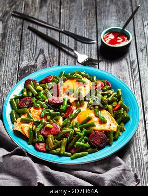 Spanish breakfast of green beans fried sunny side up eggs and spicy fried chorizo sausage on a blue plate with cutlery on a wooden table, top view, cl Stock Photo