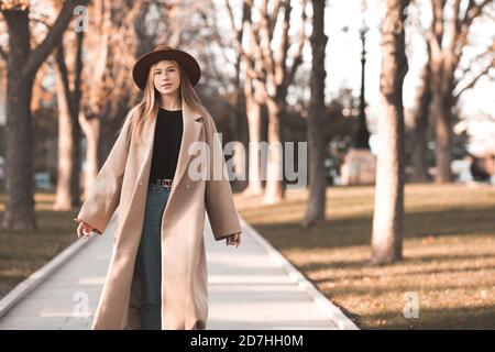 Stylish teen girl 13-14 year old wearing trendy beige coat and hat walking in autumn park. Fall season. Teenagerhood. Stock Photo