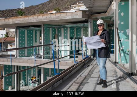Young architect wearing a safety helmet and sunglasses reading the floor plans at a construction site, professional woman looking over the house plans Stock Photo