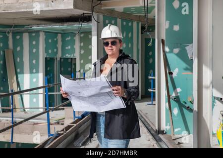 Young architect wearing a safety helmet and sunglasses reading the floor plans at a construction site, professional woman looking over the plans Stock Photo
